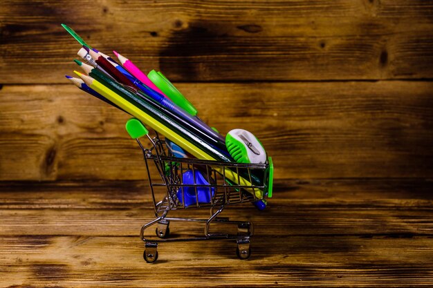 Carrito de la compra pequeño con material escolar diferente sobre fondo de madera rústica. Concepto de regreso a la escuela