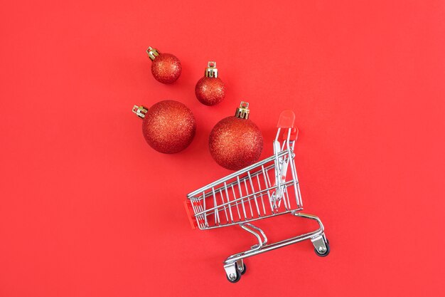 Carrito de supermercado y bolas de Navidad sobre un fondo rojo. Minimalismo, espacio de copia, plano.