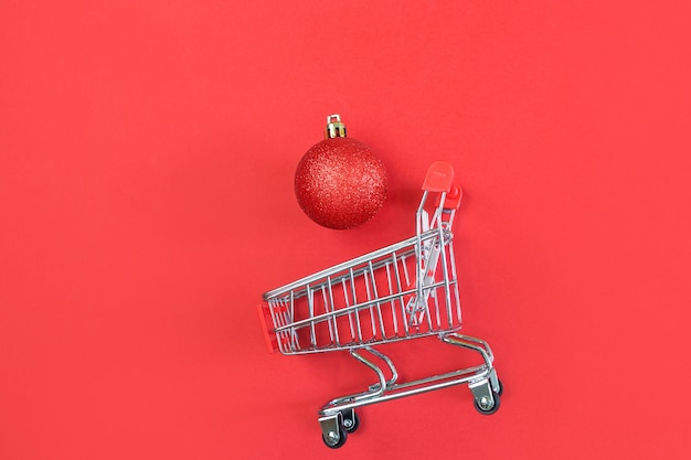 Carrito de supermercado y bolas de Navidad sobre un fondo rojo. Minimalismo, espacio de copia, plano.