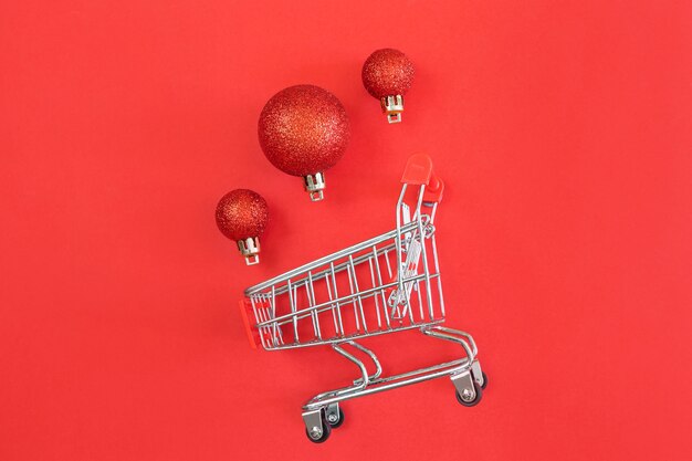 Carrito de supermercado y bolas de Navidad sobre un fondo rojo. Minimalismo, espacio de copia, plano.