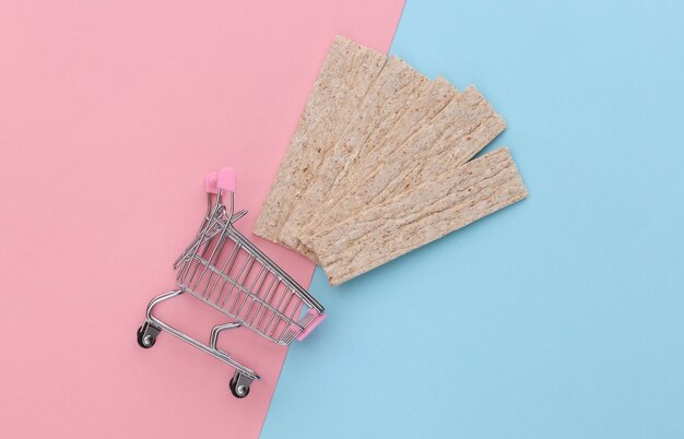 Carrito de supermercado con pan crujiente dietético sobre fondo azul pastel rosa.