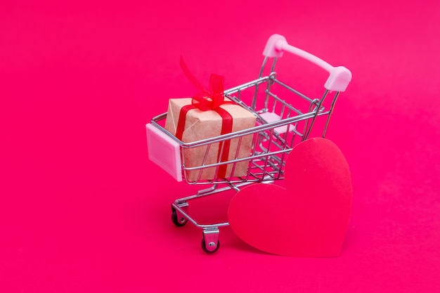 Carrito de supermercado pequeño con cajas de regalo en superficie rojo-rosa. Regala con amor el día de San Valentín