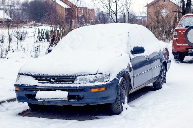 Carro coberto de neve perto de casas no inverno, após uma nevasca. Mau tempo no inverno