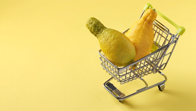 Carro de la compra con dos limones feos en mesa de color amarillo brillante. Concepto de compras ambientales, alimentos orgánicos