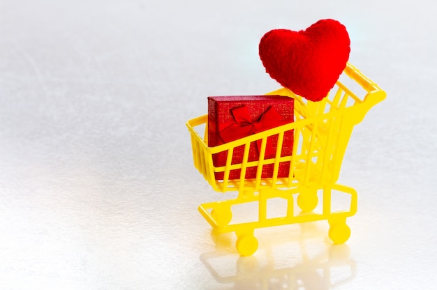 Carro de compras con caja de regalo roja y corazones rojos de confeti sobre un fondo gris copia espacio