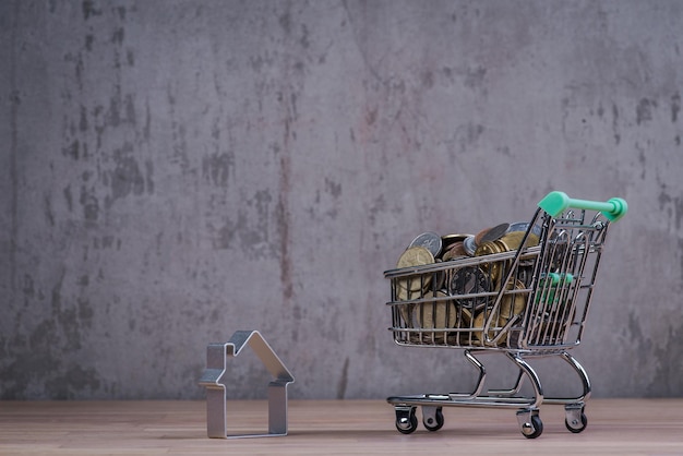 Carro de compras con casa y monedas en la mesa de madera