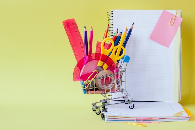 Carro de compras con diferentes artículos de papelería sobre fondo amarillo