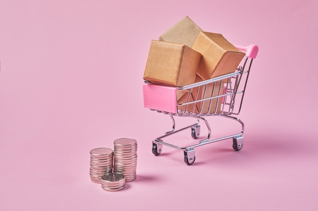 Carro de compras con paquetes de cajas y monedas sobre un fondo rosa. Carro de la tienda lleno de paquetes.