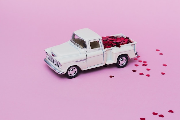 Carro de brinquedo em miniatura entregando confetes de corações vermelhos no dia dos namorados em fundo rosa