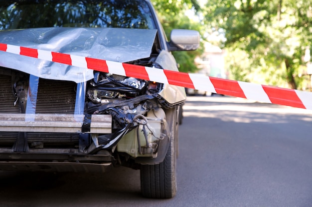 Carro destruído em acidente de trânsito acidente de carro na estrada da cidade com fita vermelha da polícia de advertência. Farol dianteiro quebrado quebrado, capô amassado sem pára-choque em acidente de carro cinza.