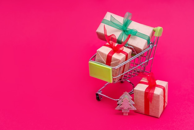 Carro de supermercado pequeño con cajas de regalo de Navidad en superficie rosa