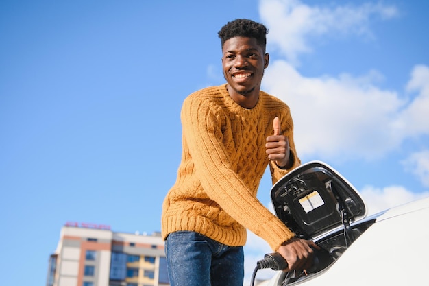 Carros elétricos, conceito EV, combustível ecológico. Retrato de jovem negro sorridente, recarregando seu carro elétrico de luxo moderno