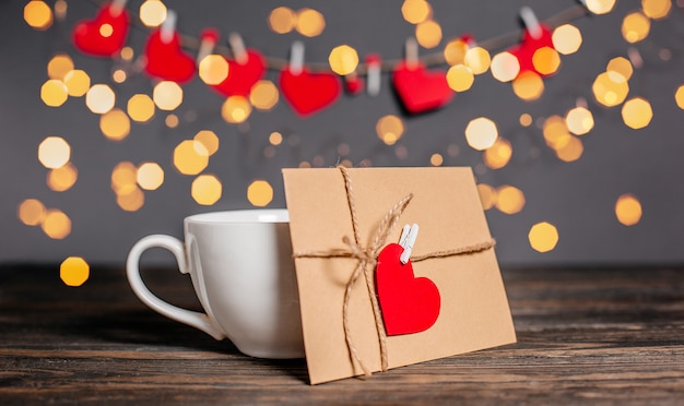 Carta de amor con un corazón junto a una taza sobre un fondo de luces, amor y concepto de San Valentín en una mesa de madera