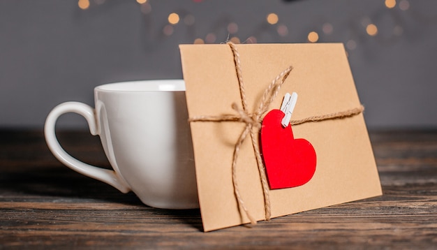 Carta de amor con un corazón junto a una taza sobre un fondo de luces, amor y concepto de San Valentín en una mesa de madera