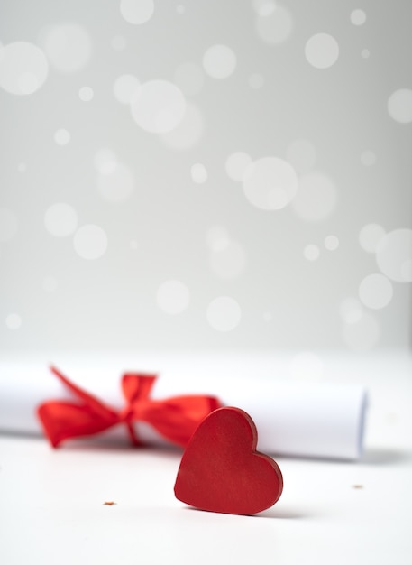 Carta de amor enrollada del día de San Valentín atada con una cinta roja y un corazón de madera rojo en el fondo bokeh claro