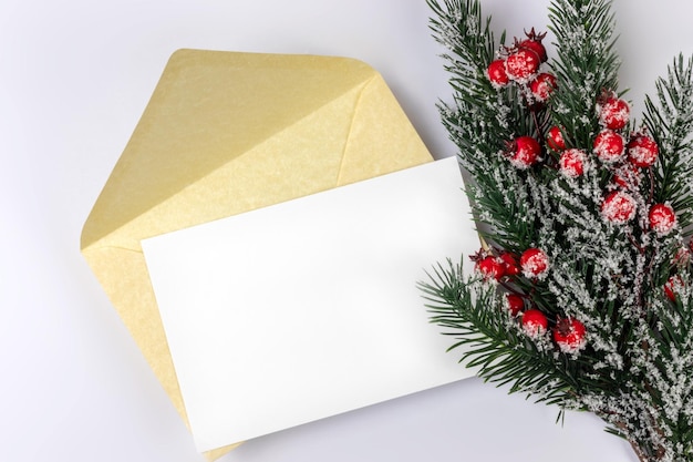 Carta de natal simulada com uma folha de papel ou cartão branco em branco vazio e um envelope dourado sobre fundo branco e um galho de árvore de natal e espaço de cópia de vista superior de azevinho