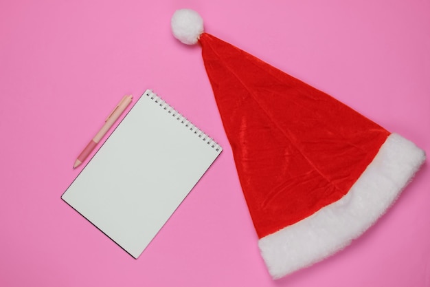 Carta a santa, lista de deseos. Gorro de Papá Noel, cuaderno con bolígrafo sobre fondo rosa. Vacaciones navideñas. Vista superior