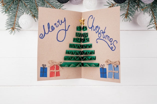 Cartão artesanal cartão com árvore de natal 3d feito de papel colorido dobrado acordeão, com a inscrição feliz natal em fundo branco com ramos de pinheiro. vista do topo