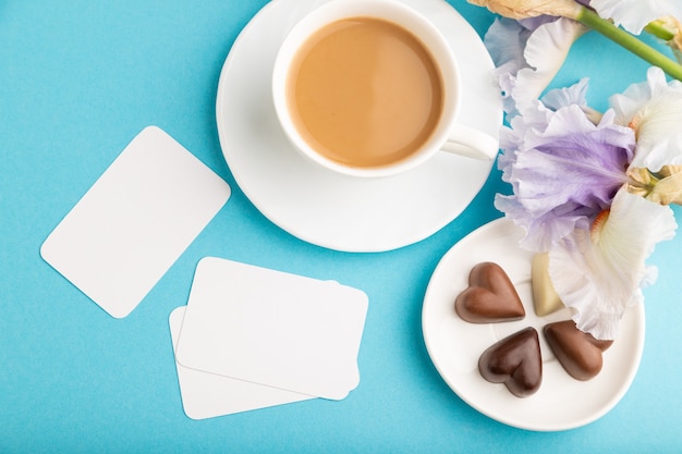 cartão branco com uma xícara de café, bombons de chocolate e flores de íris sobre fundo azul.