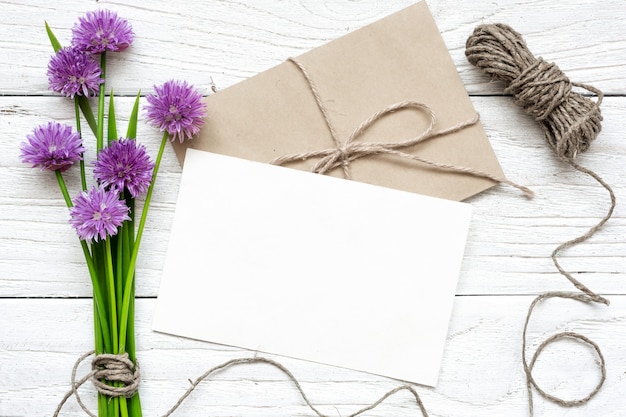 Cartão branco em branco com buquê de flores silvestres roxos e envelope com uma corda sobre a mesa de madeira branca