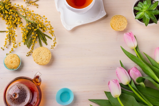 Cartão da mola com tulipas, mimosa, chá e queques na tabela de madeira.