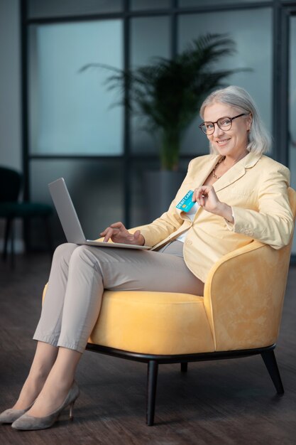 Cartão de crédito. Mulher sorridente de cabelos grisalhos em roupas elegantes, sentada em uma confortável poltrona amarela com um laptop e um cartão de débito