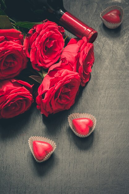 Cartão de dia dos namorados com rosas vermelhas românticas, garrafa de vinho, coração doce na mesa de madeira.