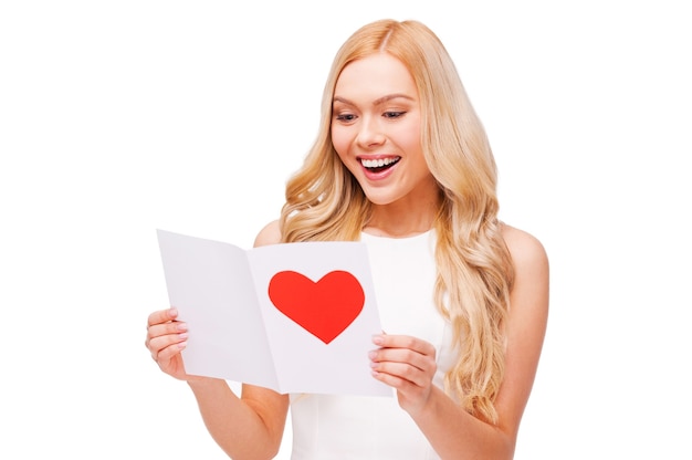 Cartão de dia dos namorados dele. Mulher jovem e bonita de cabelo loiro segurando um cartão de dia dos namorados e sorrindo em pé, isolado no branco