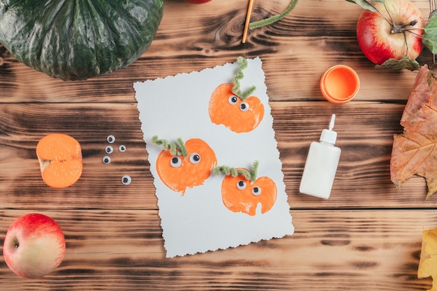 Cartão de halloween acabado feito com estampas de maçã-abóbora ao lado de cola e tinta na superfície de madeira, vista superior
