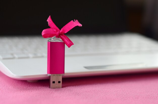 Cartão de memória flash usb rosa brilhante com um laço rosa encontra-se em um cobertor de tecido de lã rosa claro macio e peludo ao lado de um laptop branco.