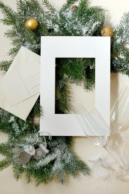 Cartão de Natal ou ano novo com galhos de árvore do abeto com bola dourada de Natal, taças de champanhe, moldura de papel branco, envelope postal. Luz dourada do bokeh. Fundo bege. Postura plana, copie o espaço