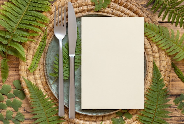 Cartão de papel em branco em um prato com faca e garfo decorado com folhas de samambaia na vista superior da mesa de madeira. Cena tropical de maquete com cartão de convite plano