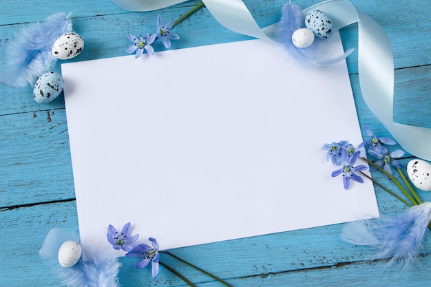 Foto cartão de páscoa branco, flores de floco de neve, ovos de páscoa e penas na placa de madeira azul.