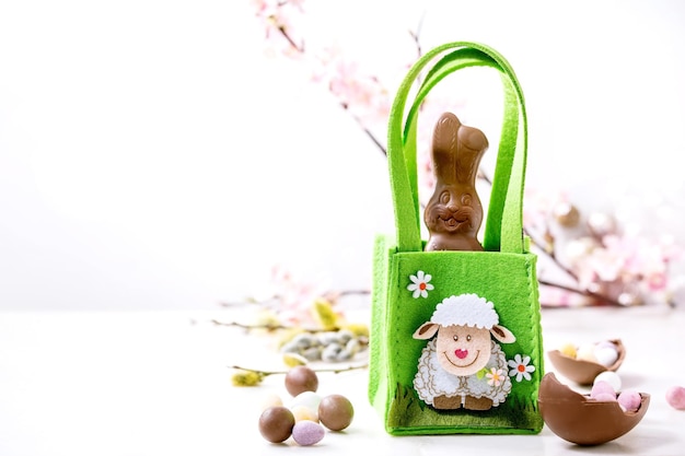 Cartão de Páscoa com coelho de doces de chocolate em saco verde, doces e ovos, ramos de salgueiro em fundo branco. Delícias de Páscoa. Copiar espaço