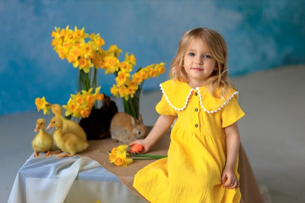 Cartão de páscoa linda garota bonita em um vestido amarelo brilhante senta-se ao lado de coelhos e patinhos perto de buquês de narcisos