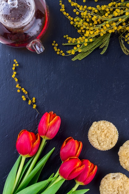 Cartão do dia das mulheres com tulipas, mimosa, chá e cupcakes no fundo da placa de pedra preta.