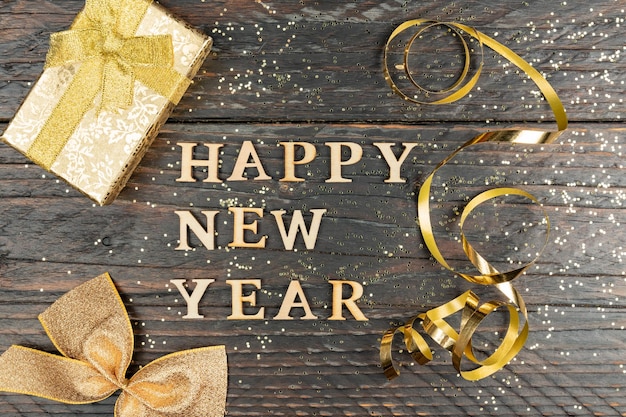 Cartão festivo com texto de feliz ano novo em bakground de madeira com confetes espumantes de ouro e caixa de presente