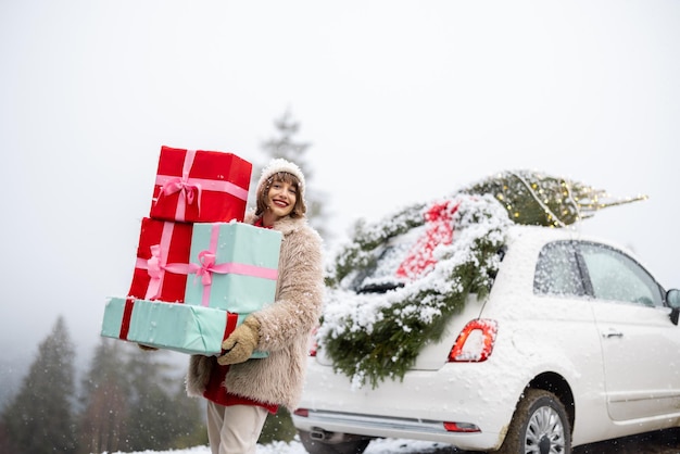 Cartão fotográfico de Natal de uma mulher com presentes e carro nas montanhas em tempo de neve