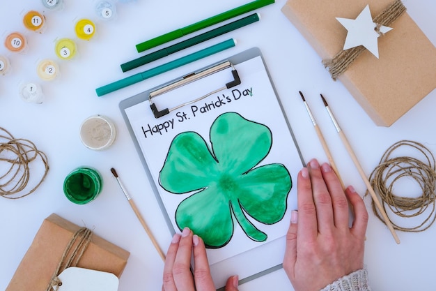 Cartão pintado caseiro dia de São Patrício. Ideia de presente, decoração. FAÇA VOCÊ MESMO. Faça Você Mesmo. Trevos, pincel e tinta. Mãos femininas. Chapéu de Saint Patricks Day.