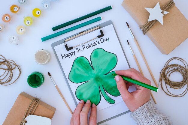 Cartão pintado caseiro dia de São Patrício. Ideia de presente, decoração. FAÇA VOCÊ MESMO. Faça Você Mesmo. Trevos, pincel e tinta. Mãos femininas. Chapéu de Saint Patricks Day.