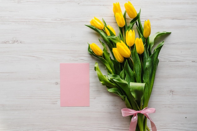 Cartão postal rosa de artesanato com buquê de tulipas amarelas em fundo de madeira Espaço para mensagem Flores de primavera Cartão de felicitações de férias para o dia das mães das mulheres dos namorados Páscoa Vista superior plana lay