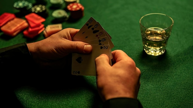 Foto cartas de pôquer nas mãos com copo de uísque e fichas de pôquer espalhadas na mesa verde