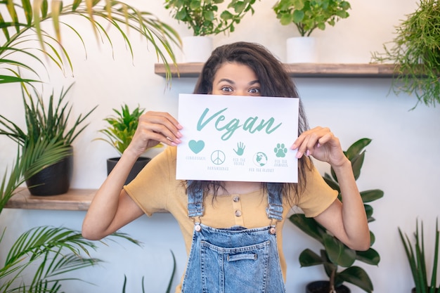 Cartaz, vegano. mulher com um pequeno pôster cobrindo a parte inferior do rosto em um ambiente fechado com plantas verdes de interior