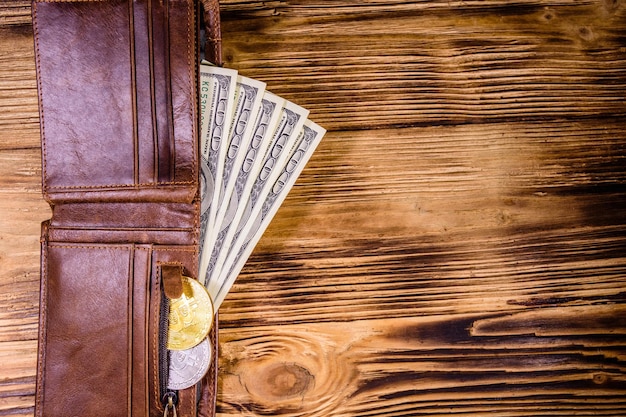 Carteira de couro marrom, bitcoins e notas de cem dólares no fundo de madeira. Vista do topo