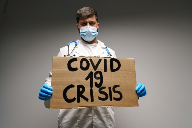 Cartel de 'Covid-19 crisis' en manos del médico manifestante sobre fondo gris.