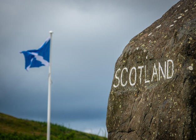 Un cartel de piedra en la frontera escocesa con una bandera de Escocia en la distancia