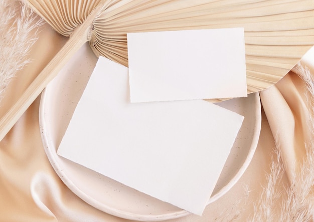 Cartões em branco no prato em folha de palmeira seca e tecido sedoso bege fecham saudação ou maquete de casamento