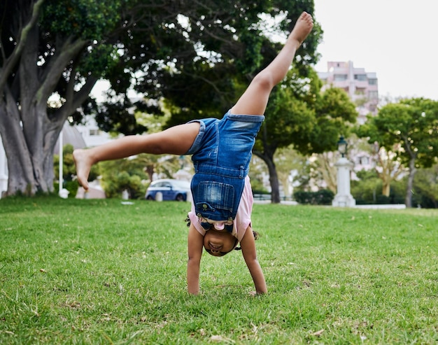 Cartwheel-Kind und Outdoor-Park Spaß eines Mädchens im Sommer mit Freiheit und Glück Lustiges fröhliches und urlaubshaftes Spiel eines jungen Kindes auf grünem Gras in der Natur an einem Morgen mit blauem Himmel und Lächeln