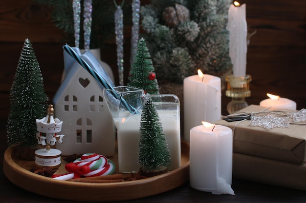 Una casa de Año Nuevo con una vela encendida dentro de decoraciones de Navidad en una bandeja de madera con velas encendidas y un vaso de leche
