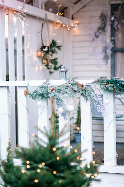 Casa blanca con porche decorado con adornos navideños y árbol de Navidad. Guirnaldas de abeto alrededor de la puerta. Hermosa terraza de invierno de la casa con guirnaldas y luces.